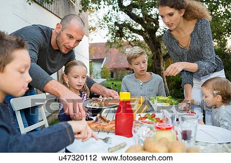 Stock Image - Father and teenage girl helping children at garden barbecue table. Fotosearch