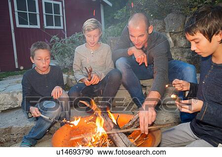 Stock Image - Father and three sons sitting by garden campfire at dusk. Fotosearch