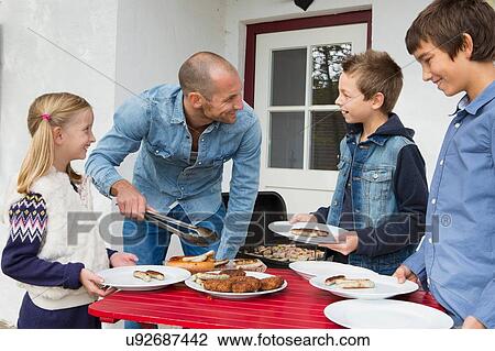 Stock Image - Father serving food to children at garden barbecue table. Fotosearch
