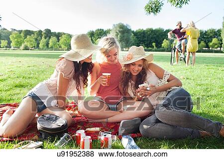 Stock Photo - Female friends reading smartphone text at park party. Fotosearch