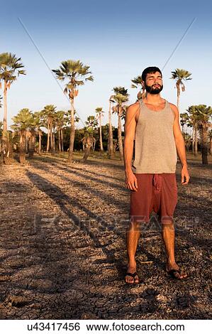 Stock Photograph - Full length front view of young man standing in front of palm trees casting shadow looking away, Taiba, Ceara, Brazil. Fotosearch