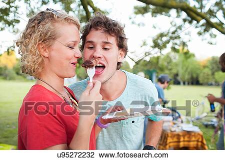 Stock Image - Happy young man and woman sharing food at sunset party in park. Fotosearch