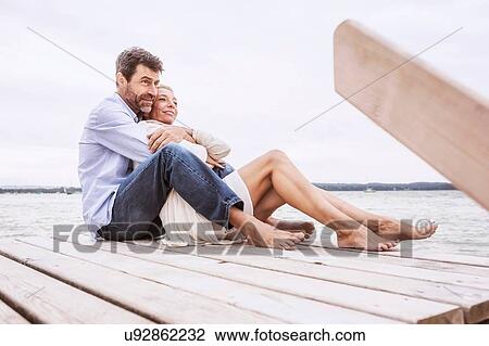 Stock Image - Mature couple sitting, hugging, on pier. Fotosearch