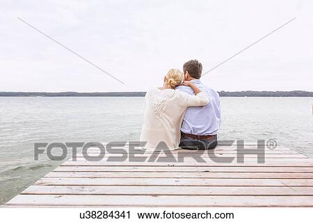 Stock Image - Mature couple sitting on pier, looking at view, rear view. Fotosearch
