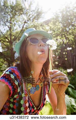 Stock Image - Mature woman wearing sunhat and sunglasses blowing dandelion seeds. Fotosearch