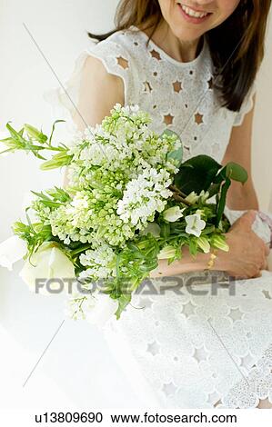 Stock Image - Mature woman wearing white broderie anglaise dress holding flower arrangement smiling. Fotosearch