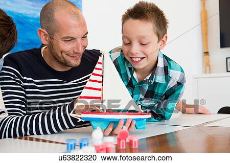 Stock Image - Mid adult man and son admiring painted toy boat at kitchen table. Fotosearch