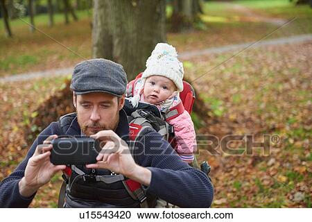 Mid adult man in park wearing flat cap carrying daughter on back in baby carrier taking selfie using smartphone View Large Photo Image Stock Image - Mid adult man in park wearing flat cap carrying daughter on back in baby carrier taking selfie using smartphone. Fotosearch
