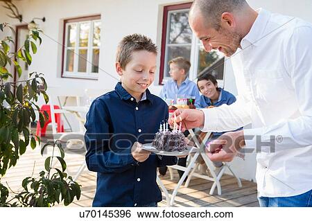 Stock Photograph - Mid adult man lighting sons birthday cake candles on patio. Fotosearch
