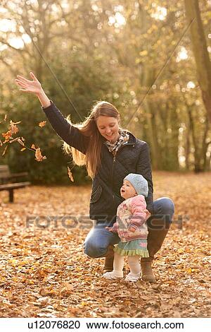 Stock Image - Mid adult woman and baby daughter watching autmn leaves in park. Fotosearch