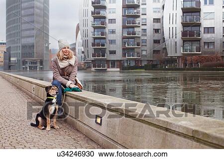 Stock Image - Mid adult woman and her dog sitting on riverside wall. Fotosearch