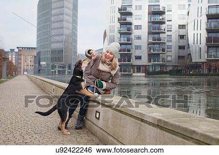 Stock Photograph - Mid adult woman feeding her dog snacks on riverside wall. Fotosearch