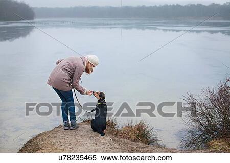 Stock Photography - Mid adult woman petting her dog on riverbank. Fotosearch