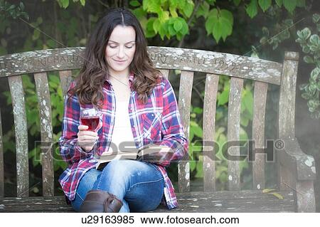 Stock Photography - Mid adult woman reading and drinking red wine on garden bench at Thornbury Castle, South Gloucestershire, UK. Fotosearch