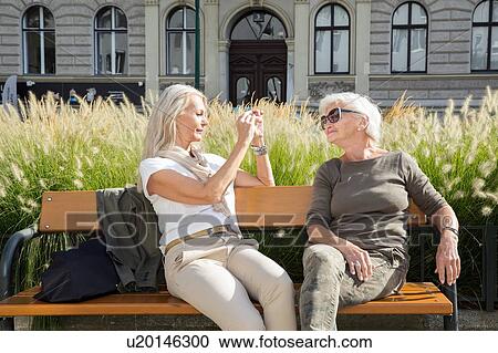 Mother and daughter sitting on bench, daughter taking photograph of mother using smartphone View Large Photo Image Stock Image - Mother and daughter sitting on bench, daughter taking photograph of mother using smartphone. Fotosearch
