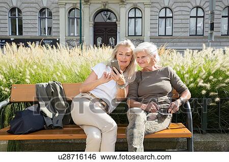 Stock Photograph - Mother and daughter sitting on bench together, looking at smartphone, outdoors,. Fotosearch