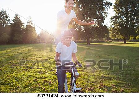 Picture - Party going man arriving in park standing on back of bicycle at sunset. Fotosearch