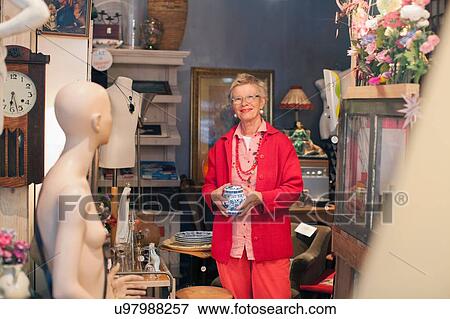 Stock Photo - Portrait of mature woman holding china pot in vintage shop. Fotosearch