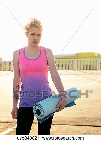Stock Photo - Portrait of young woman holding yoga mat in parking lot. Fotosearch