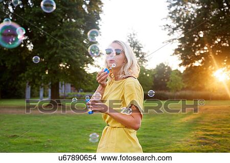 Picture - Portrait of young woman wearing yellow dress blowing bubbles in park at sunset. Fotosearch