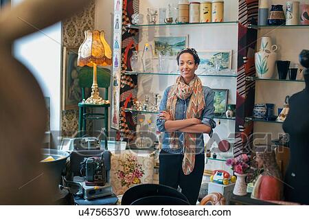 Stock Image - Portrait of young woman with arms folded in vintage shop. Fotosearch