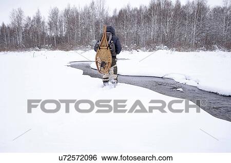 Stock Photograph - Rear view of mid adult man in snow covered field by river carrying traditional snowshoes, Ural, Russia. Fotosearch