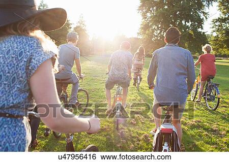 Stock Photograph - Rear view of party going adults arriving in park on bicycles at sunset. Fotosearch