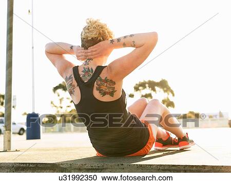 Stock Image - Rear view of young woman doing sit ups in parking lot. Fotosearch