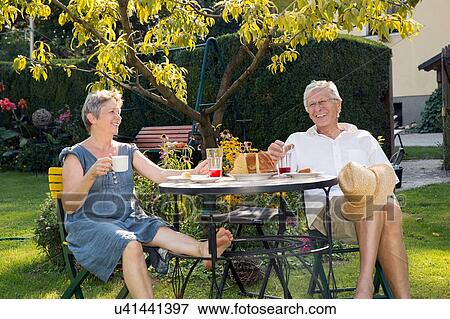 Stock Photo - Senior couple, sitting at table in garden, enjoying drink. Fotosearch