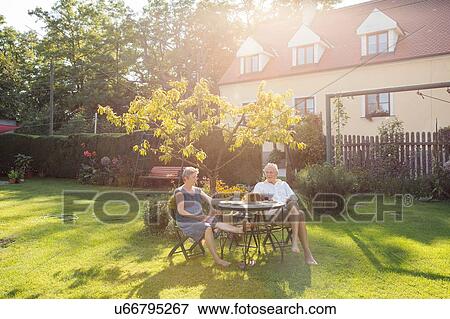 Stock Photo - Senior couple, sitting at table in garden, enjoying drink. Fotosearch