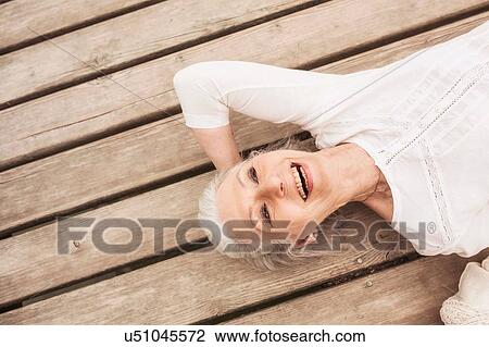 Stock Image - Senior woman relaxing on pier, smiling, elevated view. Fotosearch