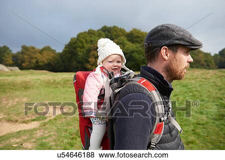Stock Photo - Side view of mid adult man in field wearing flat cap carrying daughter on back in baby carrier. Fotosearch