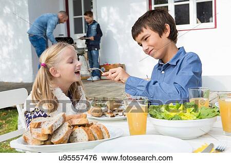 Stock Photograph - Teenage boy feeding sister at garden barbecue table. Fotosearch