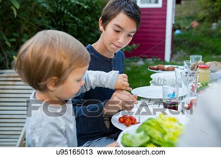 Teenage boy helping female toddler eat food at garden barbecue View Large Photo Image Stock Image - Teenage boy helping female toddler eat food at garden barbecue. Fotosearch