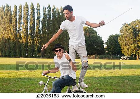 Stock Image - Two male friends fooling around on bicycle in park. Fotosearch