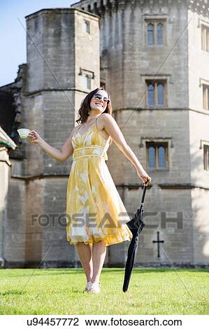 Stock Image - Woman posing with cup of tea and umbrella at Thornbury Castle, South Gloucestershire, UK. Fotosearch