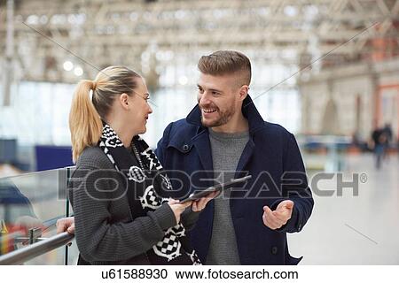 Stock Image - Young couple at train station, using digital tablet, smiling. Fotosearch