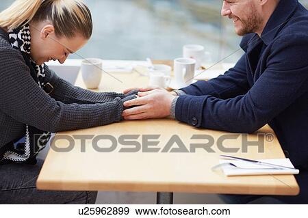 Stock Photo - Young couple sitting at cafe table, holding hands, laughing. Fotosearch