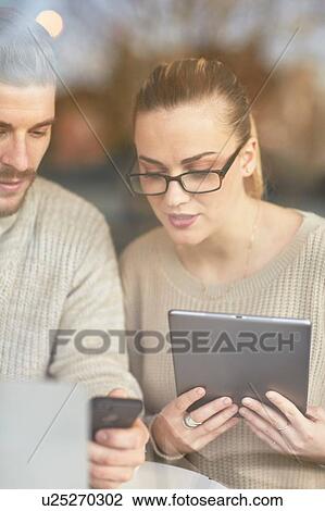 Young man and woman sitting at table, using digital tablet and smartphone View Large Photo Image Stock Image - Young man and woman sitting at table, using digital tablet and smartphone. Fotosearch