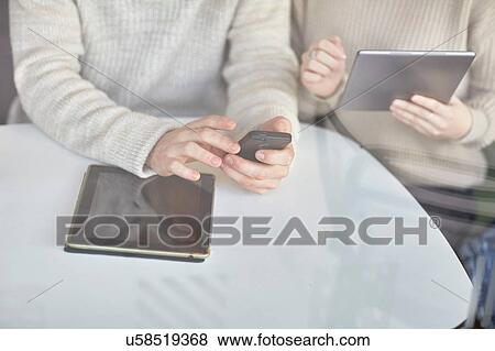 Young man and woman sitting at table, using digital tablet and smartphone, mid section View Large Photo Image Stock Photo - Young man and woman sitting at table, using digital tablet and smartphone, mid section. Fotosearch