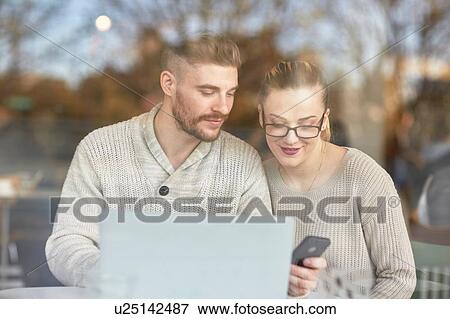 Stock Photo - Young man and woman sitting in cafe, using laptop and smartphone. Fotosearch