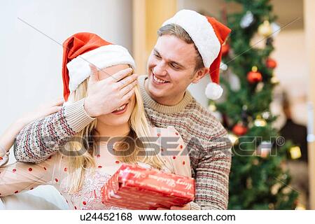 Stock Photo - Young man covering girlfriends eyes whilst handing christmas present. Fotosearch