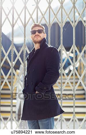 Stock Image - Young man standing by fence at train station. Fotosearch