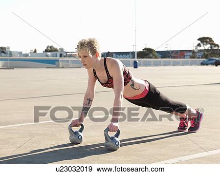 Stock Photo - Young woman doing push ups on kettlebells in parking lot. Fotosearch