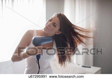 Stock Image - Young woman holding hair-dryer, drying hair. Fotosearch