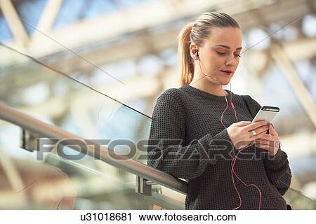 Young woman leaning against railing, wearing earphones, holding smartphone View Large Photo Image Stock Image - Young woman leaning against railing, wearing earphones, holding smartphone. Fotosearch