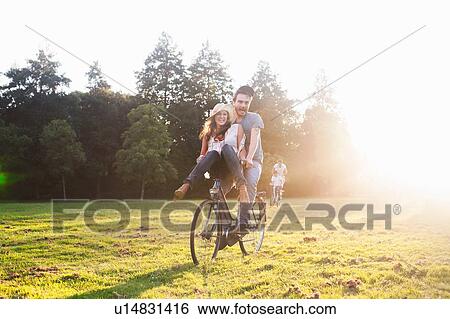 Stock Photograph - Young woman on handlebars of boyfriends bicycle at party in park. Fotosearch