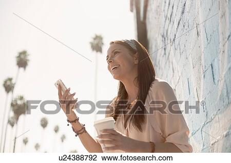 Young woman outdoors, holding coffee, using smartphone, low angle view View Large Photo Image Stock Image - Young woman outdoors, holding coffee, using smartphone, low angle view. Fotosearch