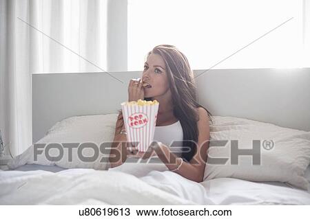 Young woman sitting in bed, eating popcorn View Large Photo Image Stock Image - Young woman sitting in bed, eating popcorn. Fotosearch