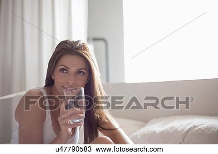Stock Image - Young woman sitting on bed, drinking glass of water. Fotosearch